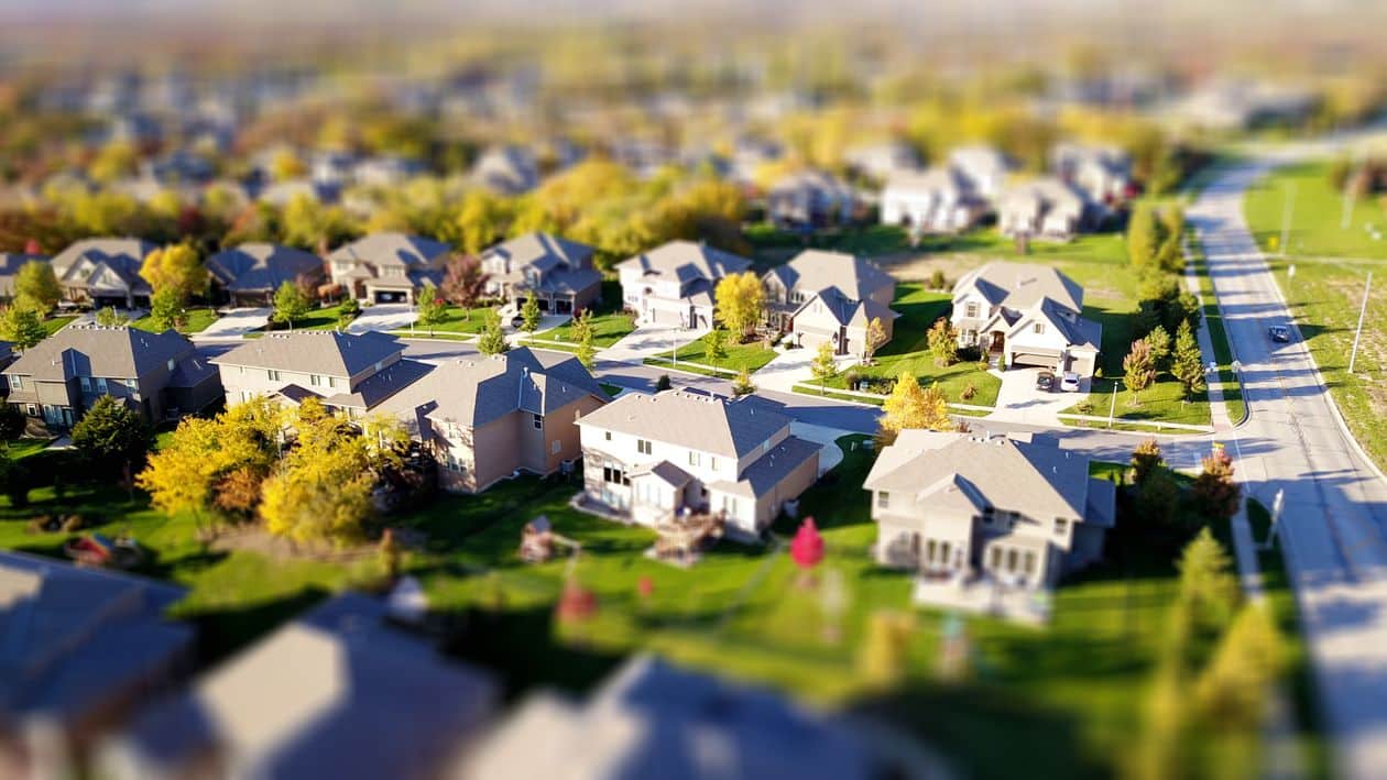 A professionally dressed real estate agent walks with purpose through a modern residential neighborhood, embodying the forward momentum and journey from classroom to closing table.